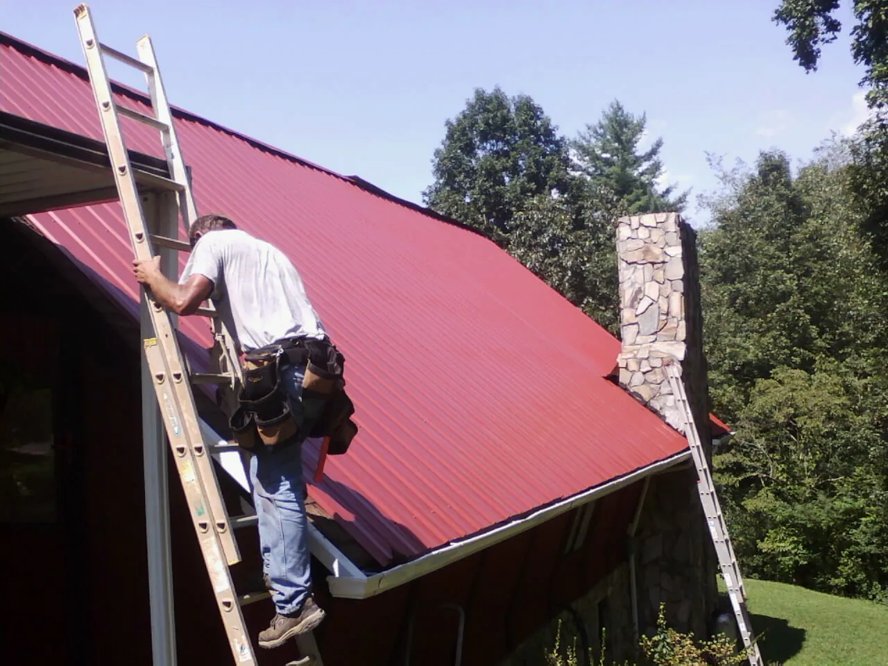 Roofer on a ladder beside a red metal roof and stone chimney — Soffit & Fascia Repair by Roofing Brothers in Lone Wolf